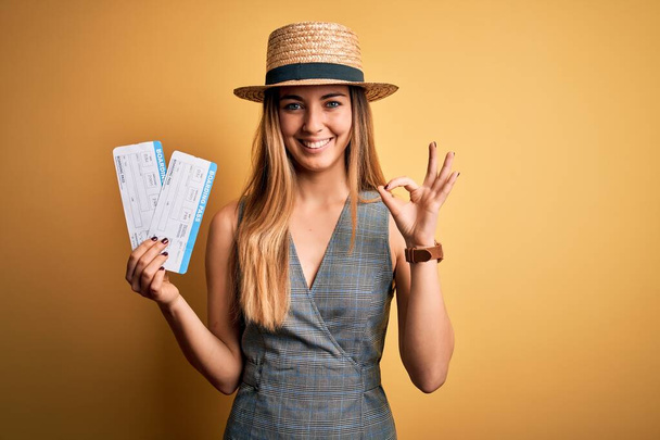 Young blonde tourist woman with blue eyes on vacation wearing hat holding boarding pass doing ok sign with fingers, excellent symbol - Foto, immagini