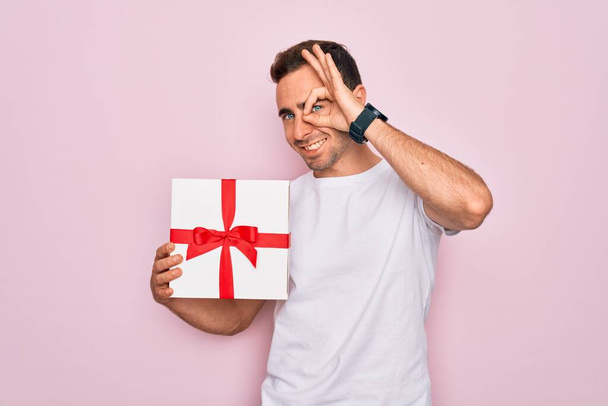 Junge gutaussehende Mann mit blauen Augen hält Geburtstagsgeschenk über isolierten rosa Hintergrund mit glücklichem Gesicht lächelnd tun ok Zeichen mit Hand auf Auge Blick durch Finger - Foto, Bild
