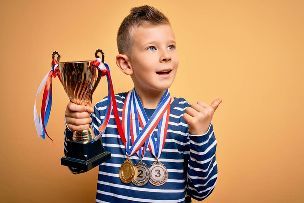 Fiatal kis kaukázusi gyerek visel győztes kitüntetések és győzelem trófea felett sárga háttér mutatva, és mutatja hüvelykujjával oldalra boldog arc mosolygós - Fotó, kép