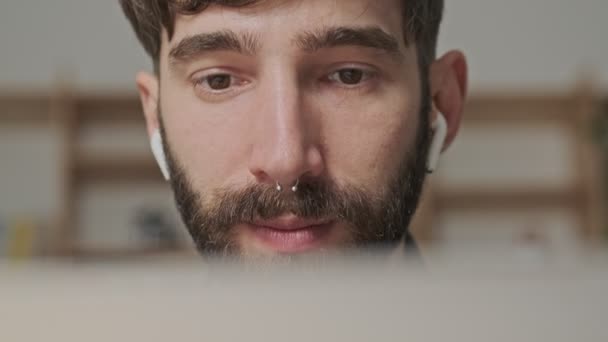 A close-up view of a thinking young digital artist in earbuds in casual clothes is looking to his laptop computer sitting in the office - Video, Çekim