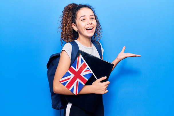 Krásné dítě dívka s kudrnaté vlasy na sobě student batoh drží pořadač a uk vlajka slaví vítězství se šťastným úsměvem a vítězný výraz se zdviženýma rukama  - Fotografie, Obrázek