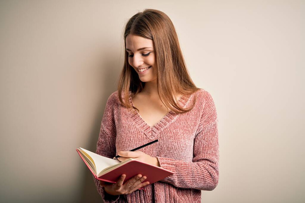 Молодая красивая рыжая студентка читает книгу на изолированном белом фоне со счастливым лицом стоя и улыбаясь с уверенной улыбкой, показывая зубы
 - Фото, изображение