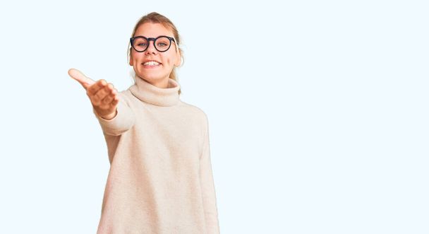 Young beautiful blonde woman wearing turtleneck sweater and glasses smiling friendly offering handshake as greeting and welcoming. successful business.  - Photo, image
