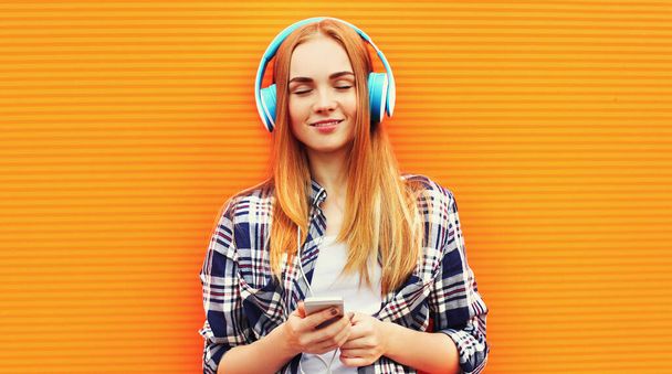 Portrait of young woman with phone in wireless headphones listening to music wearing a casual clothes on orange background - Foto, Imagem