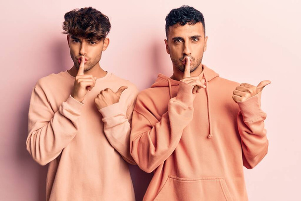 Young gay couple wearing casual clothes asking to be quiet with finger on lips pointing with hand to the side. silence and secret concept.  - Фото, изображение