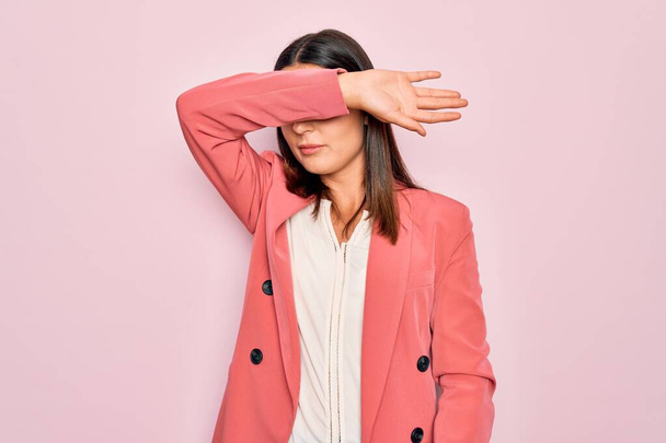 Mladá krásná brunetka byznysmena v elegantním kabátě přes izolované růžové pozadí zakrývající oči rukou, vypadá vážně a smutně. Slepý, skrývající a odmítavý koncept - Fotografie, Obrázek