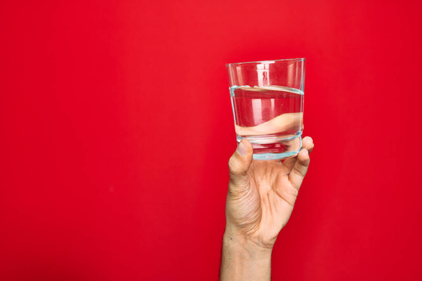 Beautiful hand of man holding glass of water over isolated red background - Foto, Imagem