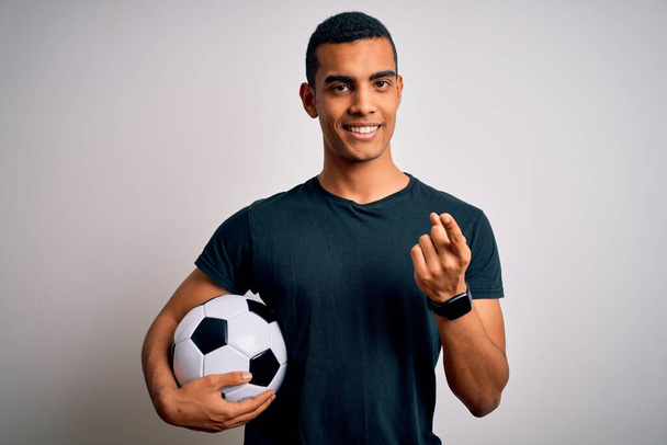 Handsome african american man playing footbal holding soccer ball over white background Beckoning come here gesture with hand inviting welcoming happy and smiling - Fotografie, Obrázek