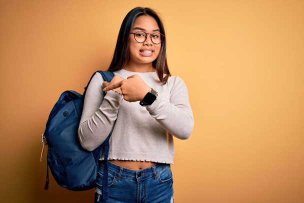 jong slim aziatische student meisje dragen rugzak staande over geïsoleerde gele achtergrond zeer gelukkig wijzend met hand en vinger - Foto, afbeelding