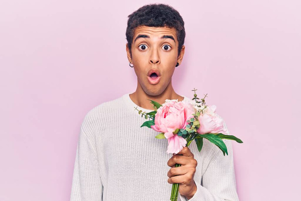 若いアフリカ系アメリカ人の男が花を持っている恐怖と驚きのために口を開けて驚いて、不信の顔  - 写真・画像