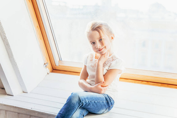 Zůstaň doma v bezpečí. Malá roztomilá roztomilá usměvavá dívka v džínách a bílém tričku sedící na parapetu v jasném světlém obývacím pokoji doma. Dětství školáci mládež relaxovat koncept - Fotografie, Obrázek
