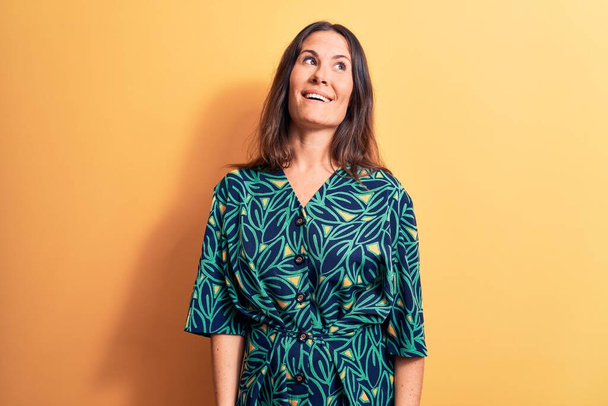 Young beautiful brunette woman wearing casual floral dress standing over yellow background looking away to side with smile on face, natural expression. Laughing confident. - Photo, Image