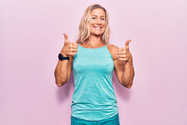 ピンクの背景にスポーツウェアを身に着けている中年の白人ブロンドの女性成功サインは、手で積極的なジェスチャーを行います,親指アップ笑顔と幸せ.陽気な表情と勝者のジェスチャー.  - 写真・画像