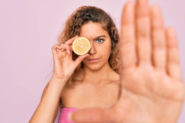 Beautiful woman with blue eyes wearing towel shower after bath holding lemon fruit over eye with open hand doing stop sign with serious and confident expression, defense gesture - Photo, Image