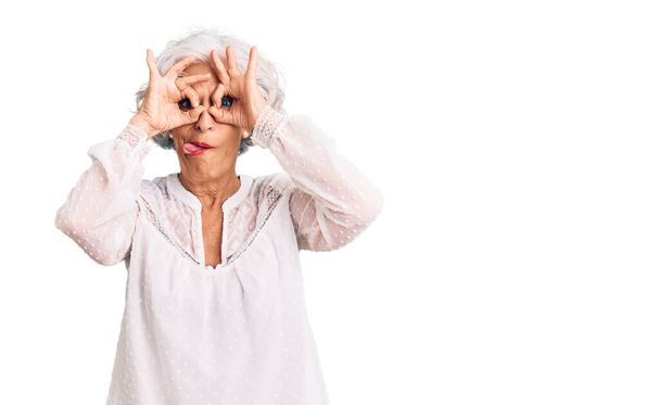 Mujer mayor de cabello gris que usa ropa casual haciendo un buen gesto como binoculares sacando la lengua, ojos mirando a través de los dedos. expresión loca.  - Foto, imagen