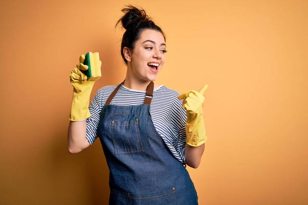 fiatal barna takarító nő visel háztartási kesztyű gazdaság súroló bozót mutatva, és mutatja hüvelykujjával oldalra boldog arc mosolygós - Fotó, kép