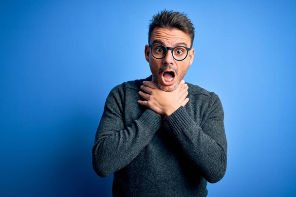 Young handsome man wearing casual sweater and glasses standing over blue background shouting and suffocate because painful strangle. Health problem. Asphyxiate and suicide concept. - Photo, Image