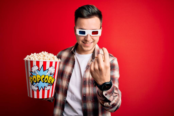 Mladý pohledný kavkazský muž v 3D filmových brýlích a jíst popcorn kývající sem přijít gesto s rukou zvoucí šťastný a usměvavý - Fotografie, Obrázek