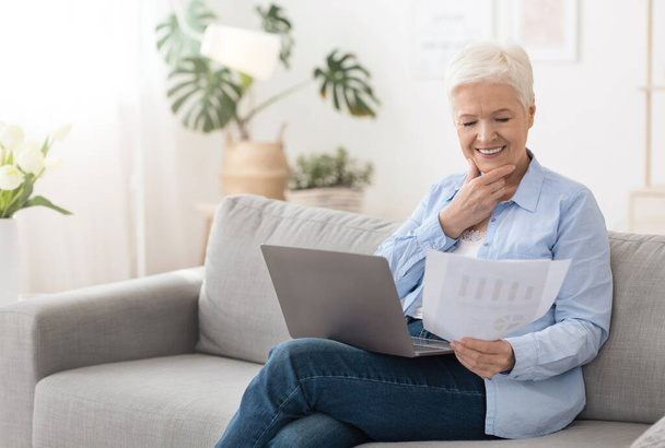 Femme âgée utilisant un ordinateur portable et lisant des documents tout en travaillant à distance à la maison - Photo, image