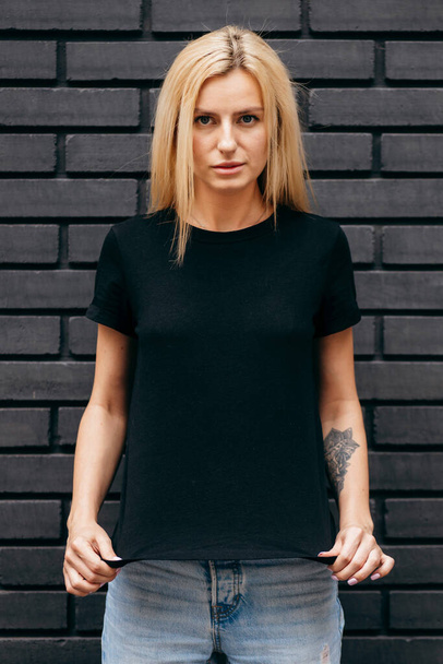 Stylish blonde girl wearing black t-shirt and glasses posing on black wall background, urban clothing style. Street photography - Photo, Image