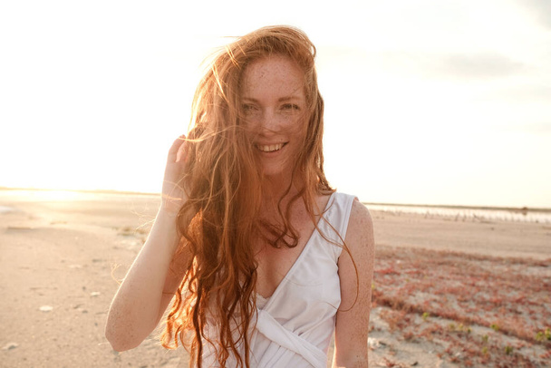Beautiful young lady with long healthy red hair and cute white dress walking near lake. Hot summer evening. Sunset. Lifestyle concept. - Valokuva, kuva
