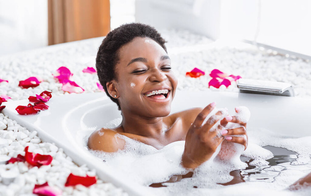Rest staying at home. Laughing african american girl in bathroom with foam and petals - Photo, Image