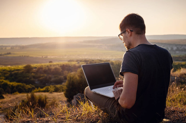 człowiek z laptopem siedzący na skraju góry z pięknym widokiem na dolinę. Koncepcja zasięgu sieci komórkowej. 5G. - Zdjęcie, obraz