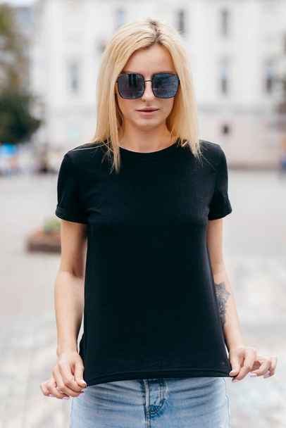 Elegante ragazza bionda che indossa t-shirt nera e occhiali in posa contro strada, stile di abbigliamento urbano. Foto di strada - Foto, immagini