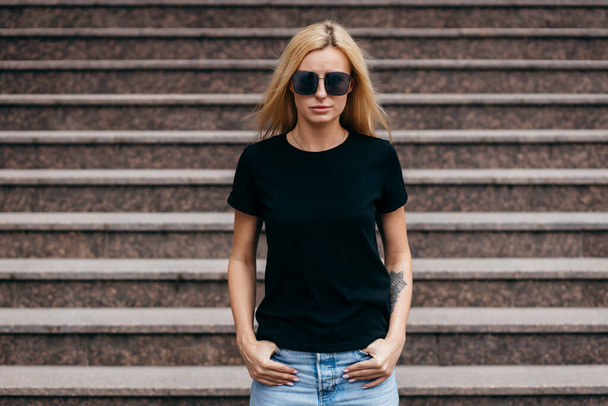 Stylowa blondynka w czarnej koszulce i okularach pozowanie przeciwko ulicy, styl odzieży miejskiej. Fotografia uliczna - Zdjęcie, obraz