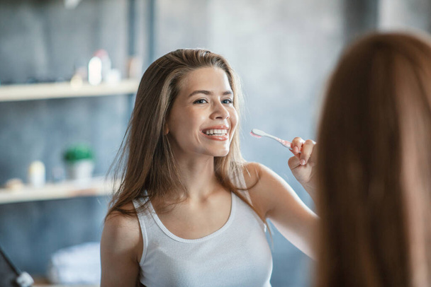 Günlük sabah rutini. Milenyum kızı diş fırçasıyla dişlerini içeride aynanın yanında temizliyor. - Fotoğraf, Görsel