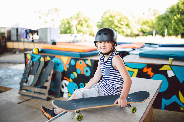 Boy sitting at skatepark, looking camera. Kid resting with skate board at skate park. child sits on ramp resting after training on skateboard. Trendy youngster enjoying free time at half pipe ramp. - Photo, Image