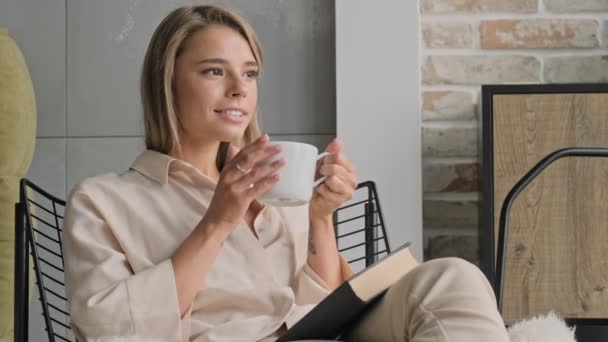 Pleased attractive blonde woman drinking tea and looking away while sitting with book on chair at home - Imágenes, Vídeo