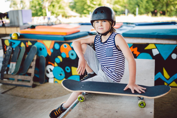 Trendy Boy Skateboarder genießen Freizeit im Skatepark, sitzt auf Halfpipe-Rampe. Ausruhen nach der Skatfahrt. Junge entspannt mit Skateboard auf Sportplatz Kind sitzt auf Rampe und wartet auf seine Freunde - Foto, Bild