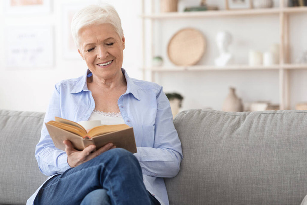 Favoriete tijdverdrijf. Glimlachende oudere dame lezen boek over comfortabele bank thuis - Foto, afbeelding