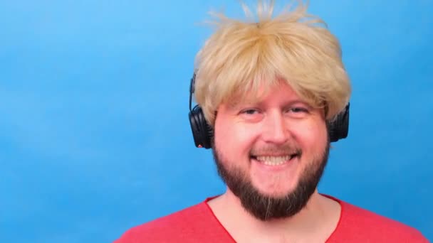 freaky fat man in a wig and a pink T-shirt in his hands listens to music on headphones and dances on a blue background - Footage, Video