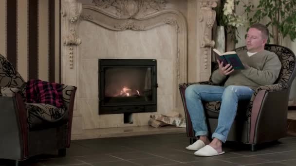 Man in chair reading book on background of fireplace. Video. Rich man reads book in armchair on background of expensive interior with fireplace. Cozy evening with book by fireplace - Footage, Video