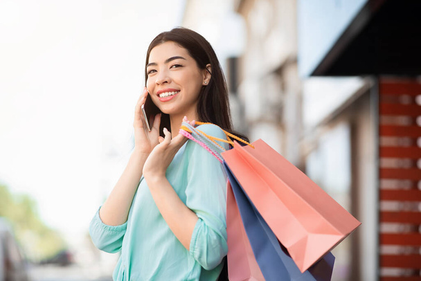 Joven mujer asiática sosteniendo bolsas de compras y hablando en el teléfono celular al aire libre
 - Foto, Imagen