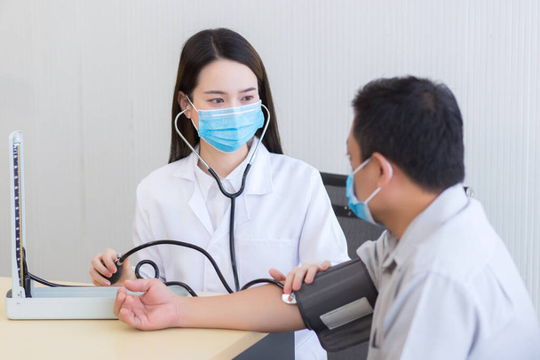 Azjatka lekarz słuchać puls (tętno) używać stetoskop zakładać na składanym stawie mężczyzna pacjent i pompować gumę miernik ciśnienia krwi. Obie noszą maski medyczne w szpitalu.. - Zdjęcie, obraz