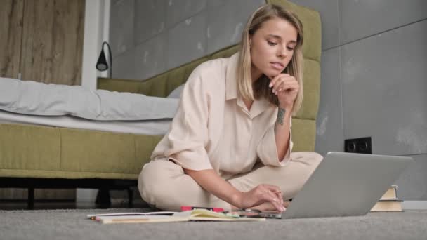 Concentrated attractive blonde woman using laptop computer while sitting on  the floor at home - Πλάνα, βίντεο