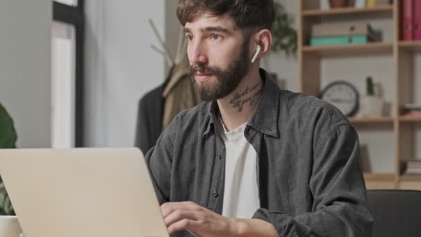A handsome young digital artist is talking on the phone using his earbuds in casual clothes while working with his silver laptop computer sitting in the office - Filmagem, Vídeo