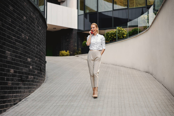 Full length of attractive blond fashionable businesswoman talking on the phone in business center exterior. - Photo, Image