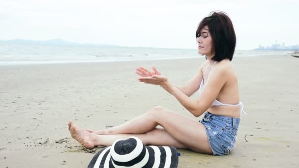 Влітку жінка-бікіні використовує сонцезахисний лосьйон spf sunblock на пляжі. Весела жінка в білому бікіні, короткі панчохи, солом'яні капелюхи, що сидять на тропічному острові з засмагою на пляжі дивіться на море
 - Кадри, відео
