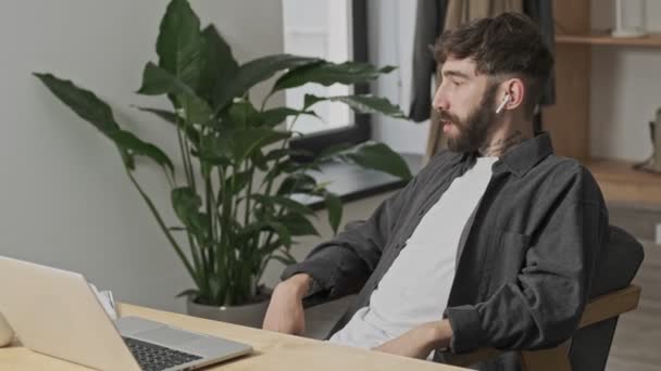 An attractive young digital artist is making notes while talking on the smartphone using his earbuds sitting in the office - Materiał filmowy, wideo