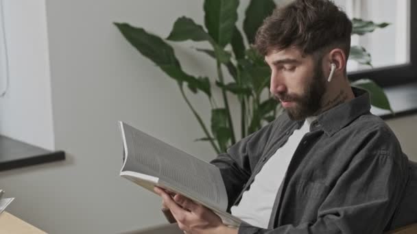 A focused young businessman wearing earbuds is reading paper book sitting in the office - Materiaali, video