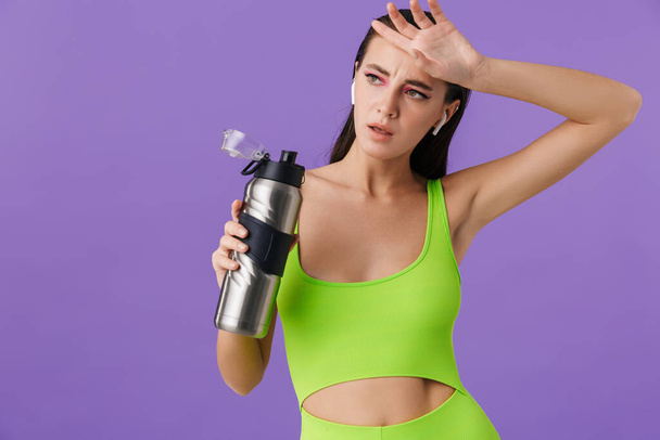 Photo of sporty tired woman with bright makeup using earpods and holding water bottle isolated over violet background - Zdjęcie, obraz
