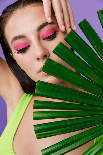 Photo closeup of young seductive woman with bright makeup holding palm leaf isolated over violet background - Photo, image