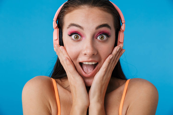 Photo closeup of alluring surprised woman using headphones and looking at camera isolated over blue background - Valokuva, kuva