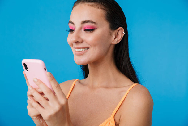 Foto close-up van verleidelijke vrolijke vrouw met behulp van mobiele telefoon en glimlachen geïsoleerd over blauwe achtergrond - Foto, afbeelding