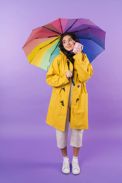 Photo of happy cheery brunette woman in yellow raincoat posing isolated over purple wall background holding umbrella and coffee. - Foto, Imagen