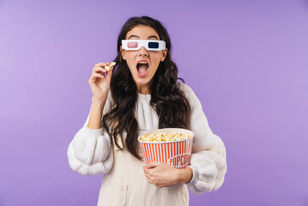 Fotografie koncentrované brunetky žena představuje izolované přes fialové stěny pozadí sledovat TV kino v 3D glsases jíst popcorn. - Fotografie, Obrázek
